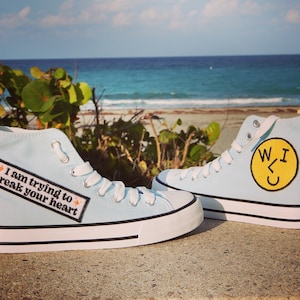 Peut inclure: Une paire de chaussures montantes en toile bleu clair avec des lacets blancs. Une chaussure a un patch smiley jaune avec le texte "W<3U" et l'autre chaussure a un patch noir avec le texte "I am trying to break your heart".