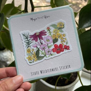 K&ouml;nnte beinhalten: Eine quadratische Tr&auml;gerkarte mit hellblauen R&auml;ndern und dem Text Maya Kiwi sowie State Wildflowers Sticker. Ein gestanzter Aufkleber in Form des Bundesstaates Oregon ist mittig platziert und zeigt eine Aquarellillustration verschiedener Wildblumen.