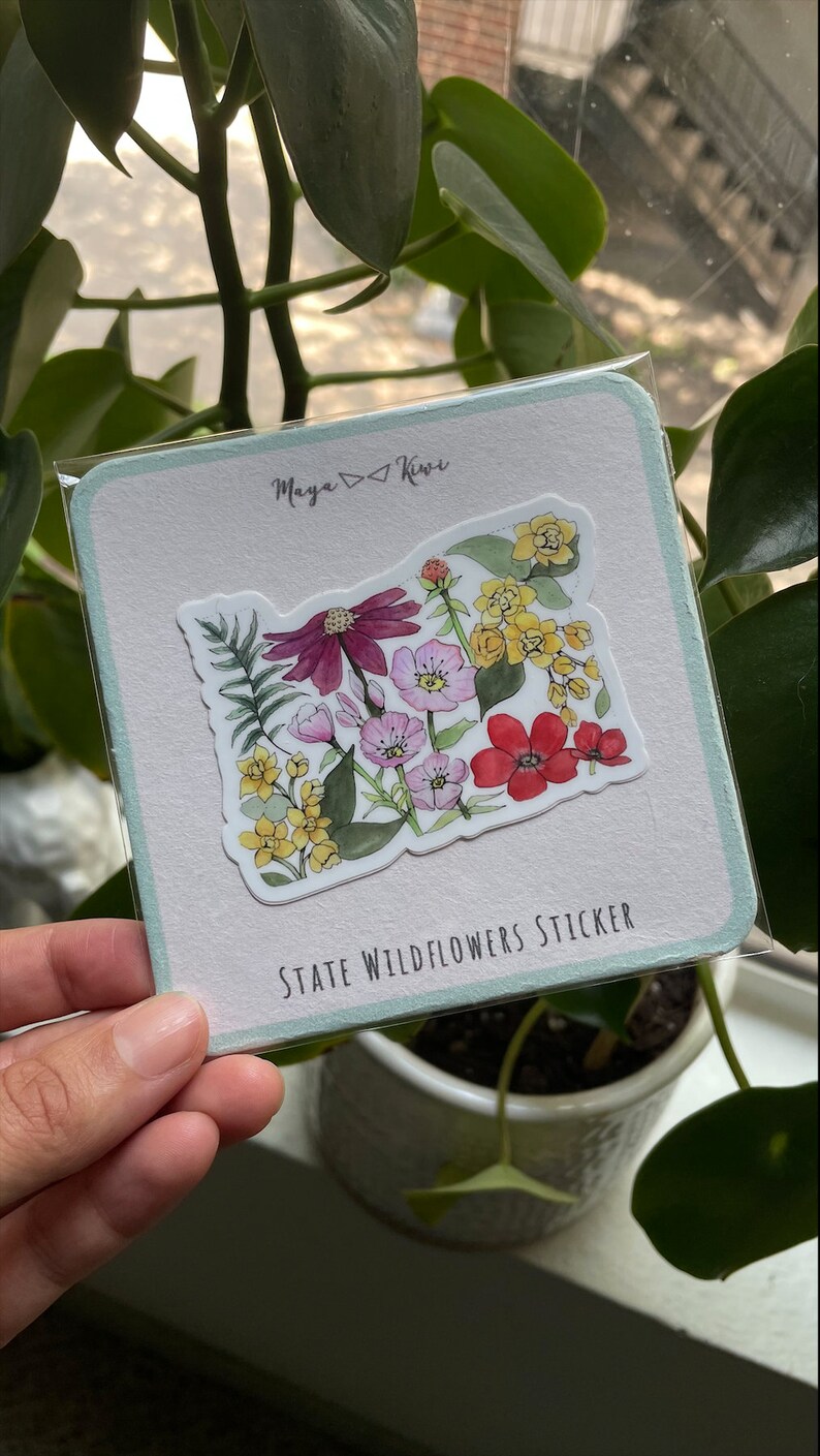 K&ouml;nnte beinhalten: Eine quadratische Tr&auml;gerkarte mit hellblauen R&auml;ndern und dem Text Maya Kiwi sowie State Wildflowers Sticker. Ein gestanzter Aufkleber in Form des Bundesstaates Oregon ist mittig platziert und zeigt eine Aquarellillustration verschiedener Wildblumen.