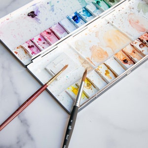 May include: A white watercolor paint palette with a variety of colors, including pink, blue, green, and yellow. The palette is open and two paintbrushes are lying next to it. One brush is brown with a gold tip, and the other is black with a gold tip.