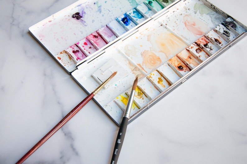 May include: A white watercolor paint palette with a variety of colors, including pink, blue, green, and yellow. The palette is open and two paintbrushes are lying next to it. One brush is brown with a gold tip, and the other is black with a gold tip.