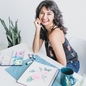 May include: A woman smiles while sitting at a table with a sketchbook, watercolor paints, and a mug. She is working on a watercolor painting of flowers.