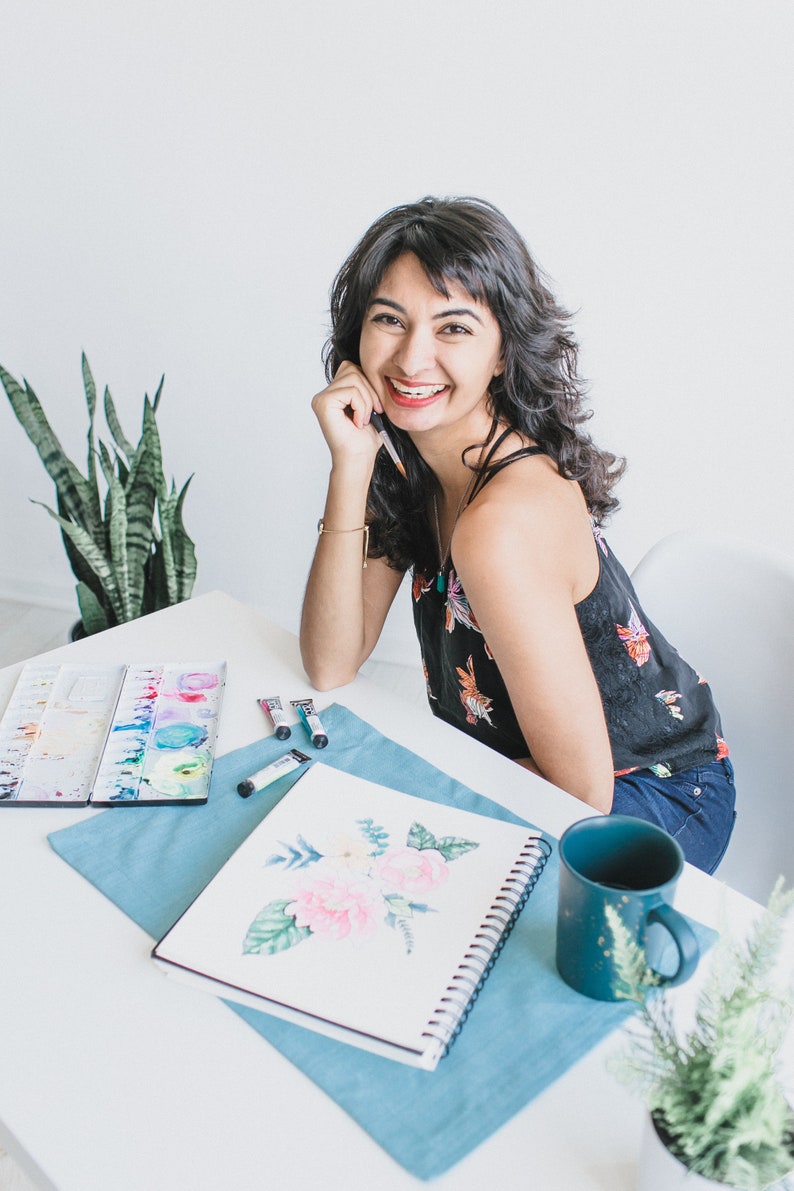 May include: A woman smiles while sitting at a table with a sketchbook, watercolor paints, and a mug. She is working on a watercolor painting of flowers.