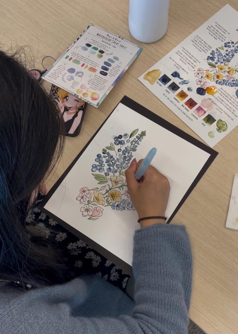 K&ouml;nnte beinhalten: Ein Aquarellbild mit blauen, gelben und rosafarbenen Blumen auf wei&szlig;em Papier, das mit einem Wassertankpinsel gemalt wird. Neben dem Papier liegen zwei informative Aquarell-Anleitungsbl&auml;tter mit Farbmustern und Text sowie ein Wasserbeh&auml;lter.