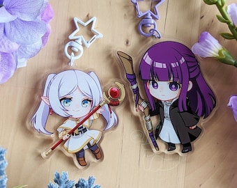 Könnte beinhalten: Zwei Acryl-Schlüsselanhänger mit Anime-Figuren. Eine Figur hat weißes Haar und ein weißes Outfit mit einem rot gekrönten Stab. Die andere hat violettes Haar und einen dunklen Mantel mit einem Holzstab. Beide haben sternförmige Verschlüsse.