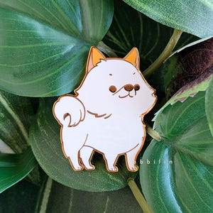 Könnte beinhalten: Weißer Emaille-Pin mit einem kleinen, flauschigen Hund mit orangefarbenen Ohren. Der Hund steht mit leicht geneigtem Kopf. Der Pin hat einen goldenen Umriss.