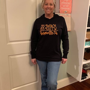 May include: A black long-sleeved shirt with a hood. The shirt has orange text that reads "35.91606°N 83.9207°W Knoxville, TN".