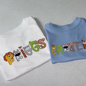 May include: Two white t-shirts with animal-themed letters spelling out the names "Briggs" and "Carter". The shirts are on a white surface.