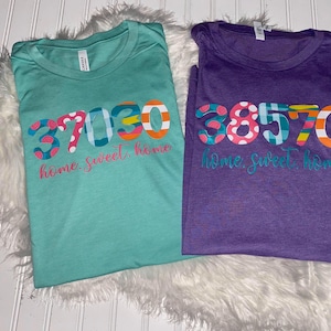May include: Two t-shirts with the text "home sweet home" and a colorful number design. The shirt on the left is teal and the shirt on the right is purple.