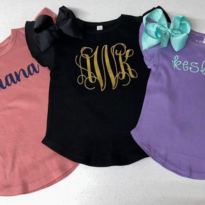 May include: Three personalized t-shirts with different names and designs. The first shirt is a dusty rose color with the name "Eliana" in navy blue lettering. The second shirt is black with a gold monogram "MVR". The third shirt is lavender with the name "Kesleigh" in light blue lettering.