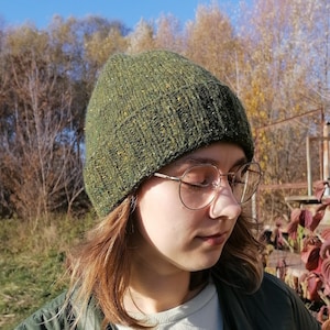 May include: A green hand knit beanie with flecks of yellow and orange. The fold-over brim of the hat is ribbed, the top part is plain.  The hat is worn with a green quilted jacket and round gold-rimmed glasses. The background features trees and a clear blue sky, suggesting an outdoor setting.