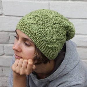 May include: A woman wearing a green knitted hat with a cable knit pattern. The hat is pulled down over her ears and she is looking down.