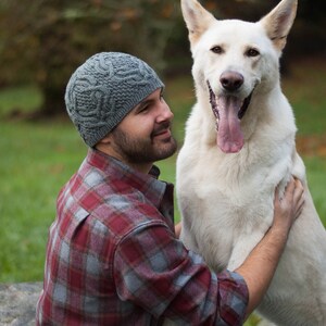 May include: A man wearing a gray knitted hat and a red and gray plaid shirt is holding a white dog with its tongue sticking out.