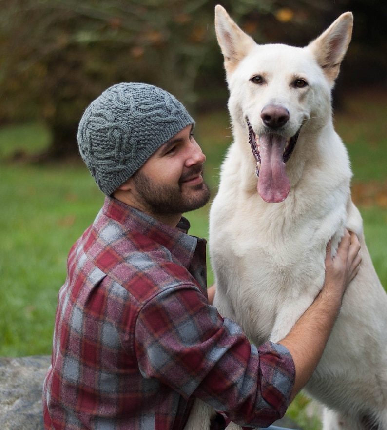 May include: A man wearing a gray knitted hat and a red and gray plaid shirt is holding a white dog with its tongue sticking out.