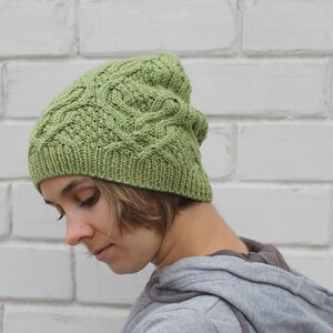 May include: A green knitted hat with a cable knit pattern. The hat is worn by a person with brown hair.