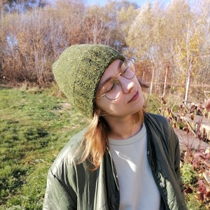 May include: A green knit beanie with a textured appearance. The beanie is worn with a green quilted jacket and round gold-rimmed glasses. The background features trees and a clear blue sky.