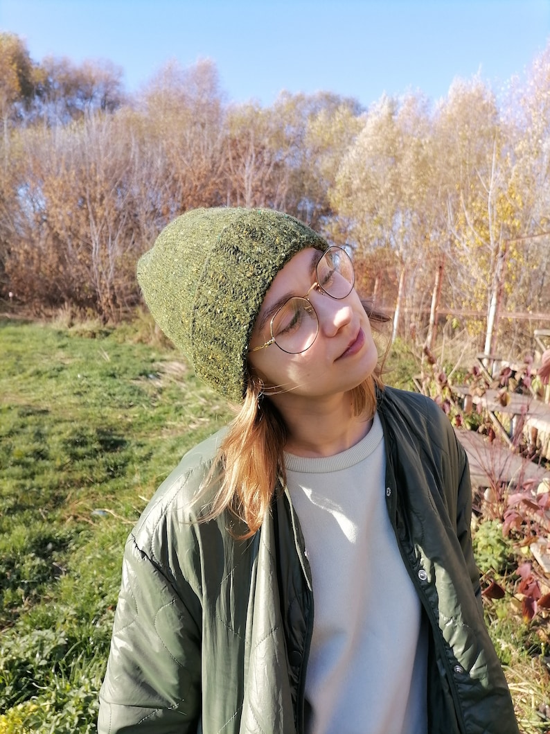 May include: A green knit beanie with a textured appearance. The beanie is worn with a green quilted jacket and round gold-rimmed glasses. The background features trees and a clear blue sky.