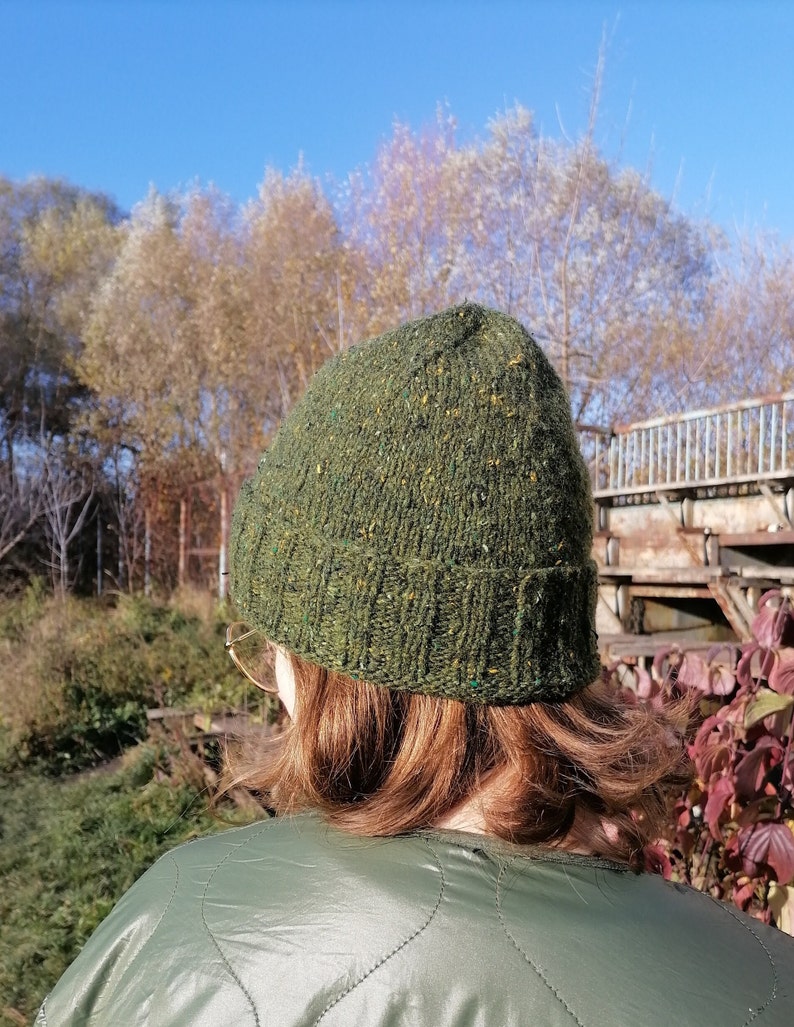 May include: A green knit beanie with flecks of color is worn by a person. The hat is made of a textured yarn and has a ribbed brim. The person is wearing a green quilted jacket. The background features trees and a blue sky.