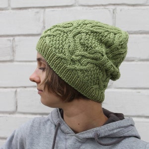 May include: A green knitted beanie with a cable knit pattern. The beanie is worn by a person with brown hair.