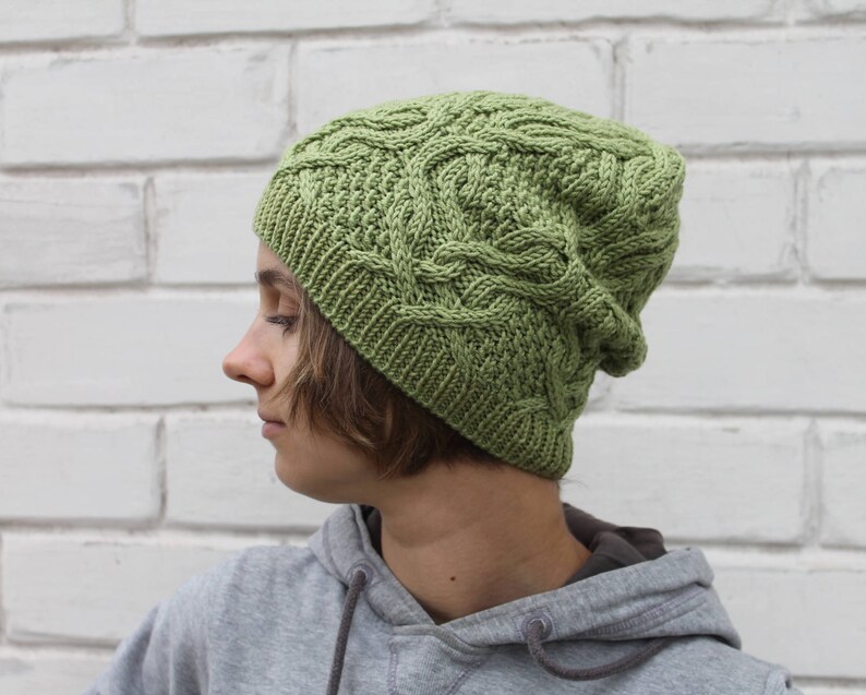 May include: A green knitted beanie with a cable knit pattern. The beanie is worn by a person with brown hair.