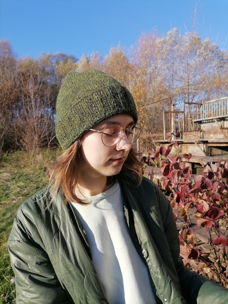 May include: A green hand knit beanie with flecks of yellow and orange. The fold-over brim of the hat is ribbed, the top part is plain.  The hat is worn with a green quilted jacket and round gold-rimmed glasses. The background features trees and a clear blue sky, suggesting an outdoor setting.