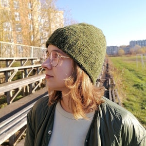 May include: A green hand knit beanie with a textured appearance in the minimalist style. The hat is worn with a green quilted jacket and round, gold-rimmed glasses. The background includes a sunny sky and a building.
