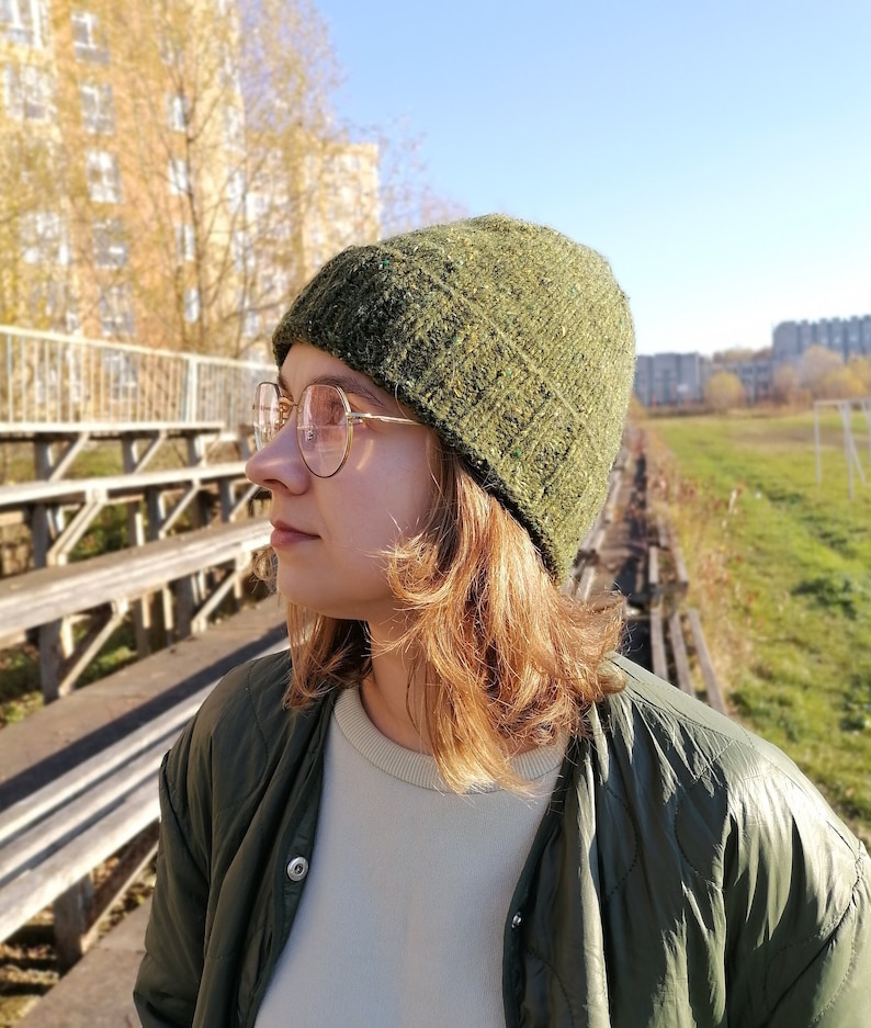 May include: A green hand knit beanie with a textured appearance in the minimalist style. The hat is worn with a green quilted jacket and round, gold-rimmed glasses. The background includes a sunny sky and a building.