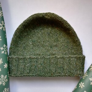 May include: A green hand knit beanie hat with a folded brim framed with snowflake wrapping paper. The hat is made of a textured yarn with flecks of color throughout. The brim is ribbed, the top of the hat is plain. The beanie is a winter accessory.