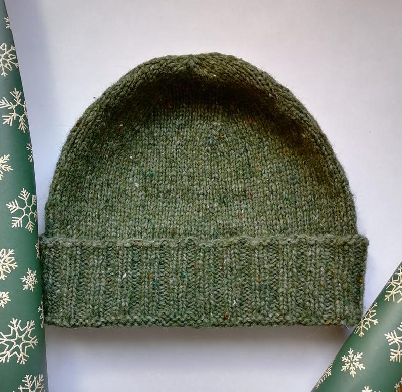 May include: A green hand knit beanie hat with a folded brim framed with snowflake wrapping paper. The hat is made of a textured yarn with flecks of color throughout. The brim is ribbed, the top of the hat is plain. The beanie is a winter accessory.