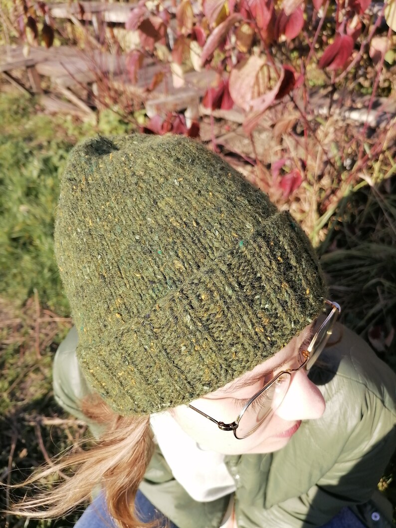 May include: A green knit beanie with flecks of color throughout. The hat is made of a textured yarn and has a folded ribbed brim. The beanie is shown from a high angle, with the top of the head and part of the face visible.