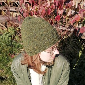 May include: A green hand knit beanie with flecks of yellow and brown. The hat is worn by a person wearing a green quilted jacket. The background features red and green autumn foliage and a wooden fence.