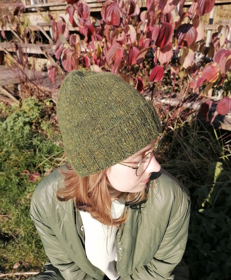 May include: A green hand knit beanie with flecks of yellow and brown. The hat is worn by a person wearing a green quilted jacket. The background features red and green autumn foliage and a wooden fence.