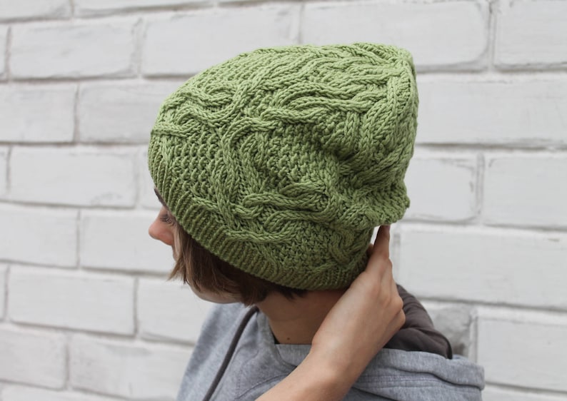 May include: A green knitted hat with a cable knit pattern. The hat is worn by a person with brown hair.