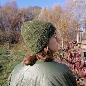 May include: A green knit beanie with flecks of yellow and brown. The hat is worn by a person wearing a green quilted jacket. The background features trees and a clear blue sky, suggesting an outdoor setting.