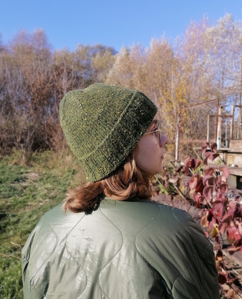 May include: A green knit beanie with flecks of yellow and brown. The hat is worn by a person wearing a green quilted jacket. The background features trees and a clear blue sky, suggesting an outdoor setting.