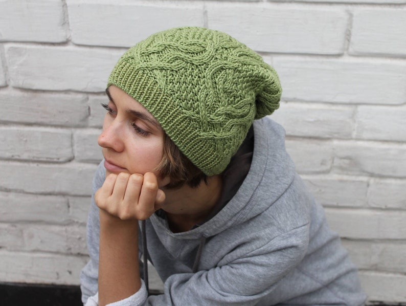 May include: A woman wearing a green knitted hat with a cable knit pattern. The hat is pulled down over her ears and she is looking down.