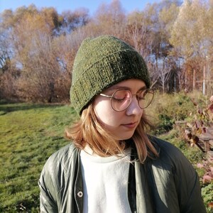 May include: A hand green knit beanie with a textured appearance. The hat is worn with a green jacket and round eyeglasses. In the background is a sunny autumn landscape: a grassy area and trees under a blue sky.