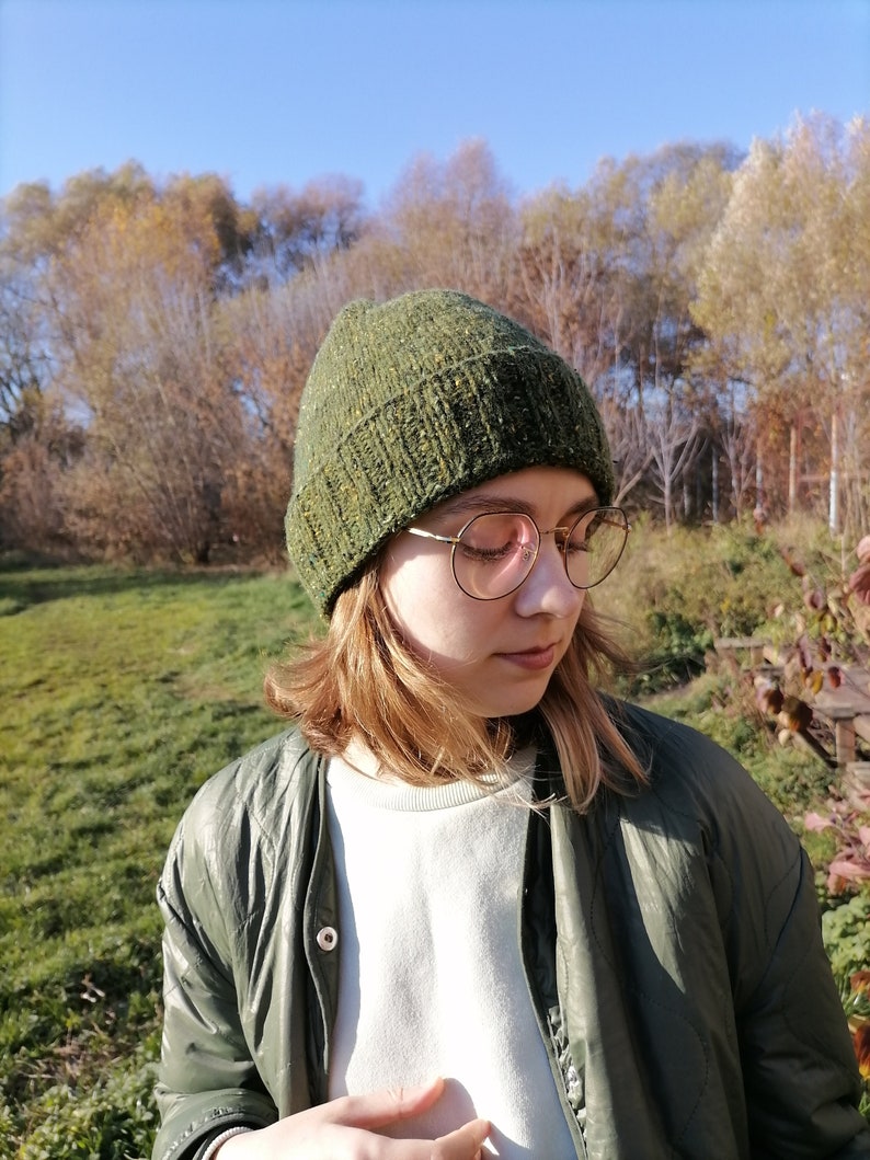 May include: A hand green knit beanie with a textured appearance. The hat is worn with a green jacket and round eyeglasses. In the background is a sunny autumn landscape: a grassy area and trees under a blue sky.