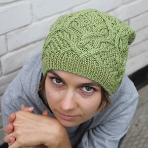 May include: A green knitted hat with a cable knit pattern. The hat is worn by a person with short brown hair and they are looking at the camera.