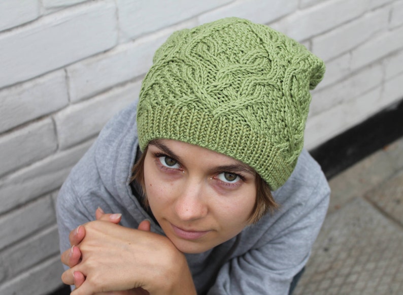 May include: A green knitted hat with a cable knit pattern. The hat is worn by a person with short brown hair and they are looking at the camera.