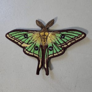 May include: An embroidered patch shaped like a moth with vibrant green wings, dark brown borders, and yellow accents near the center. The moth features two long, pointed tail-like extensions at the bottom and dark circular spots on each wing.