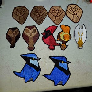 May include: A selection of embroidered bird patches. The patches depict owls in brown tones, a red cardinal, an orange bird, a white and gold bird, and two blue jays. The patches are suitable for clothing or accessories.