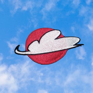 May include: Embroidered patch featuring a white cloud design over a red circle, outlined in black. The patch has a textured appearance and is set against a blue sky background with scattered clouds. The patch is likely for clothing or accessories.