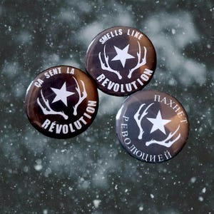 Op de afbeelding: Drie ronde zwarte en witte knoppen met een wit ster- en geweidesign. De tekst op de knoppen luidt "Smells Like Revolution", "Ca Sent La Revolution" en "Paxnet Revolyutsiey".