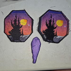 Puede incluir: Parches bordados con la silueta de un castillo contra un cielo de atardecer con una luna amarilla. Los parches son octagonales y tienen un borde negro. También hay un parche morado en forma de ataúd.