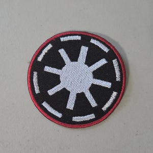 May include: A circular embroidered patch featuring a black background with a central white eight-pointed star emblem. The design includes a white dashed circular border and a thin red outer edge, creating a distinct graphic symbol.