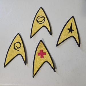 May include: Four yellow, delta-shaped embroidered patches with black borders. Each patch features a distinct black or red symbol: a black circle, a black star, a black spiral, and a red cross. These fabric emblems are arranged in a loose cluster.
