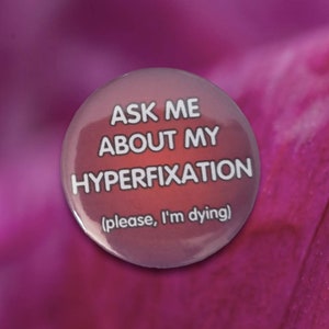 Op de afbeelding: Een roze knoop met de tekst "Ask me about my hyperfixation (please, I'm dying)"