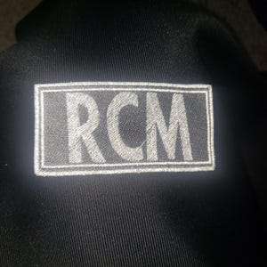 Op de afbeelding: Een zwarte patch met witte stiksels met de tekst "RCM" in hoofdletters.