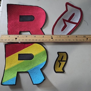 May include: Two large letter R patches, one solid red and one with diagonal rainbow stripes, alongside two smaller stylized G patches in silver and gold. A wooden ruler is placed across the items, showing the R patches are 5 inches wide.